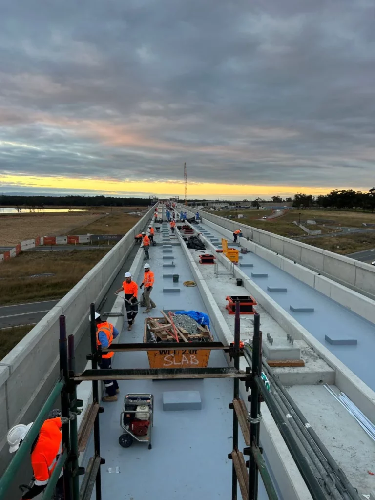 Construction workers in orange vests work on a long, elevated bridge at sunset, with equipment and materials spread along the structure under a cloudy sky.