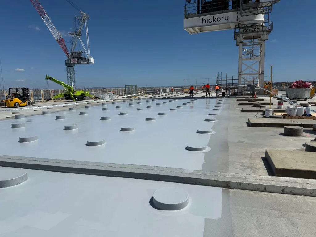 Construction site with a large flat roof covered in round capped columns, workers in safety gear, cranes, and equipment under a clear blue sky.