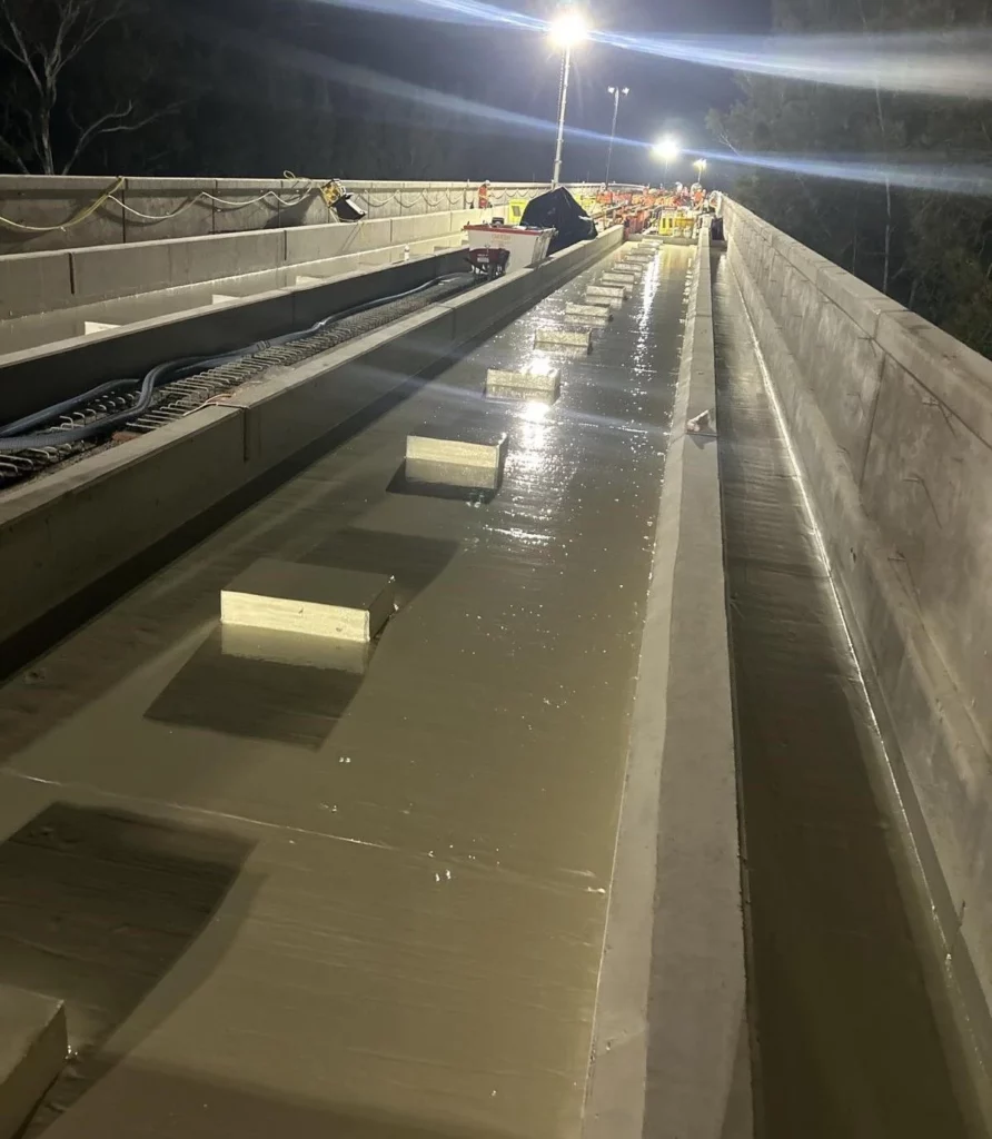 A freshly poured concrete bridge deck at night, illuminated by bright construction lights, with wooden blocks spaced evenly across the wet surface.