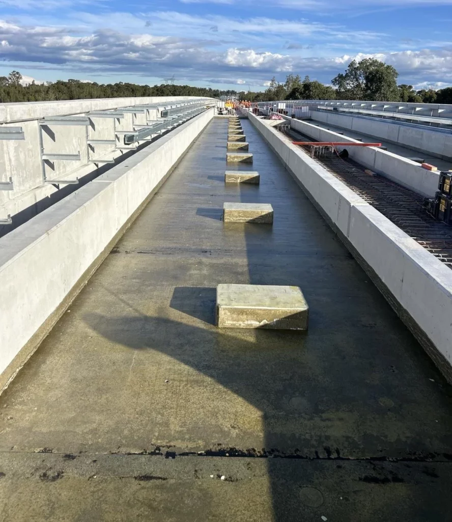 A wide concrete bridge under construction with rectangular blocks spaced along its center; railings line both sides, and construction materials are visible in the distance under a partly cloudy sky.