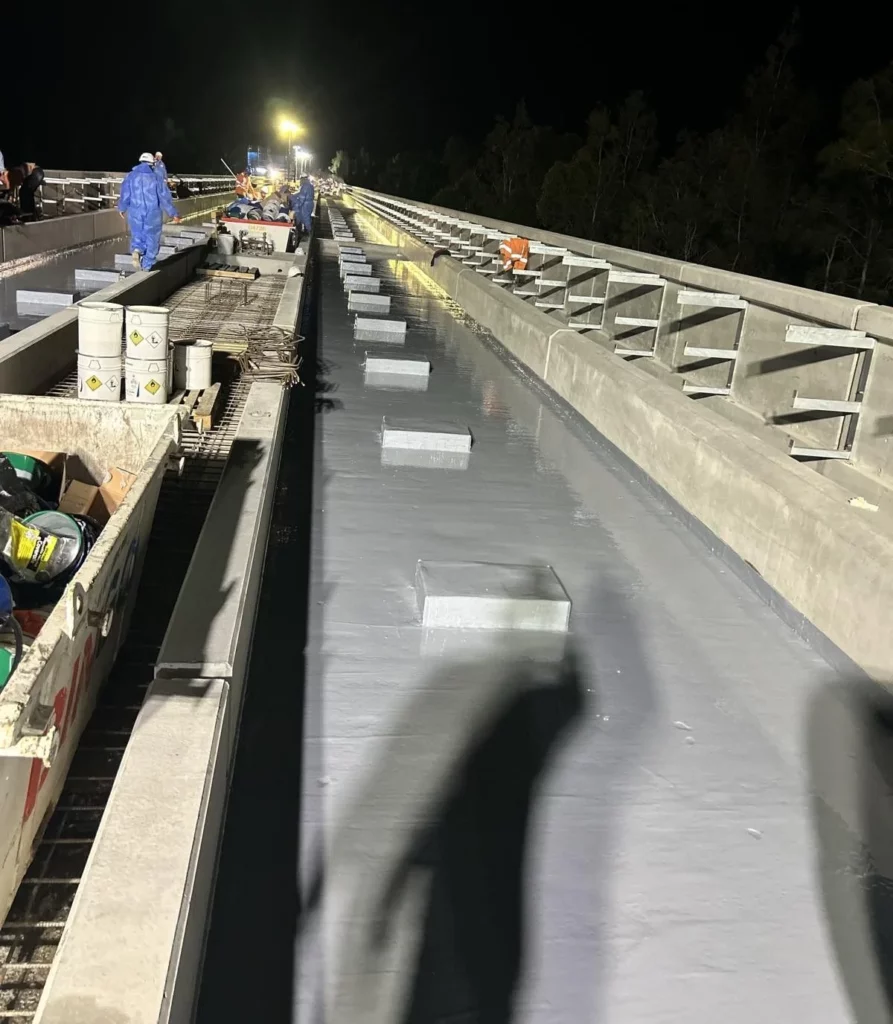 Workers in safety gear apply a waterproof membrane to a long bridge at night. Bright lights illuminate the scene, and construction materials are visible along the bridge edges. Shadows of people are cast on the surface.
