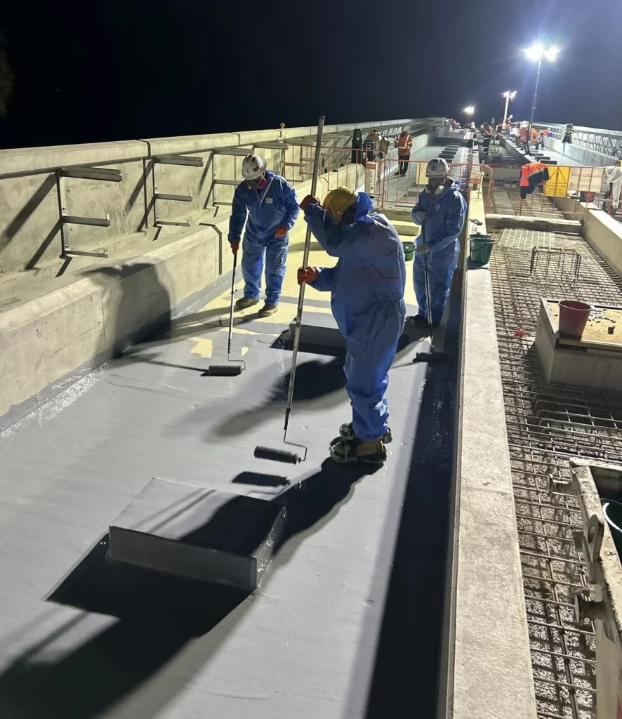Workers in blue protective suits apply coating to a bridge surface at night using long rollers. Bright lights illuminate the area, and other workers and construction materials are visible in the background.