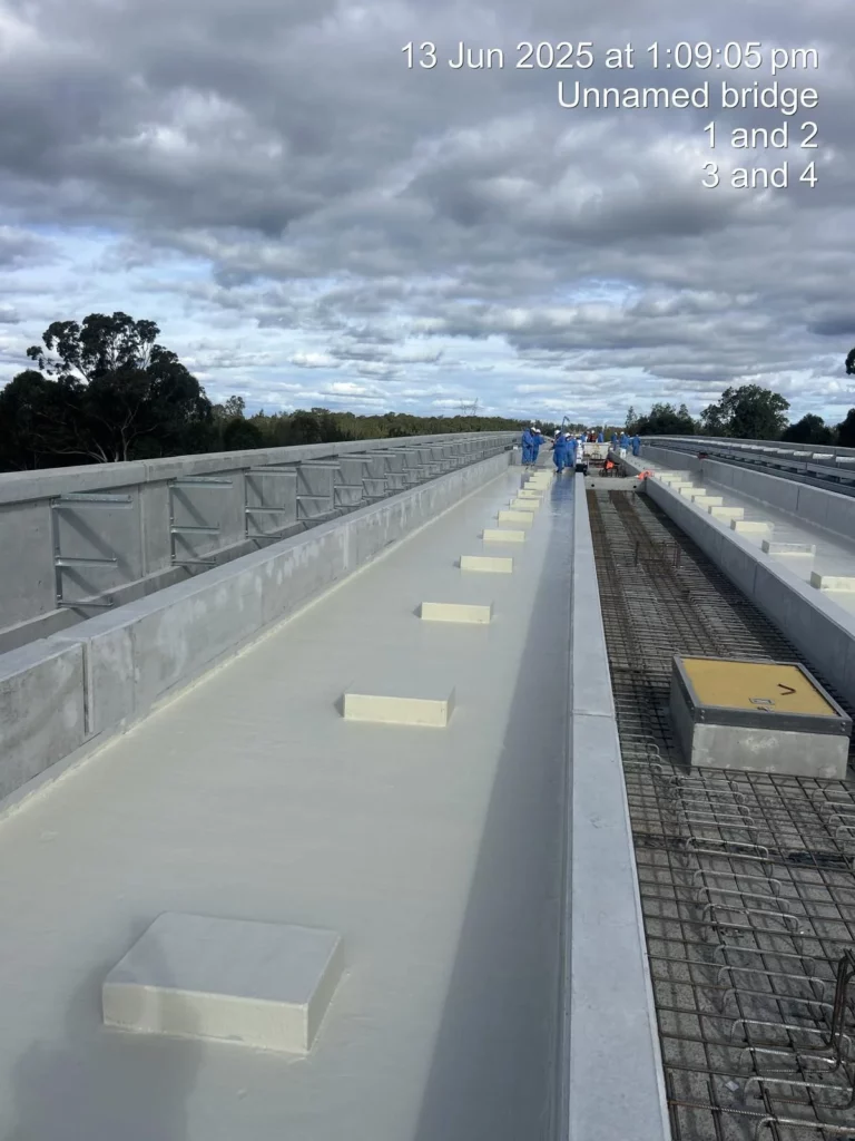 Workers in safety gear on an unfinished concrete bridge on a cloudy day; some sections are covered, others show exposed rebar. Text with date, time, and labels appears in the upper right corner.