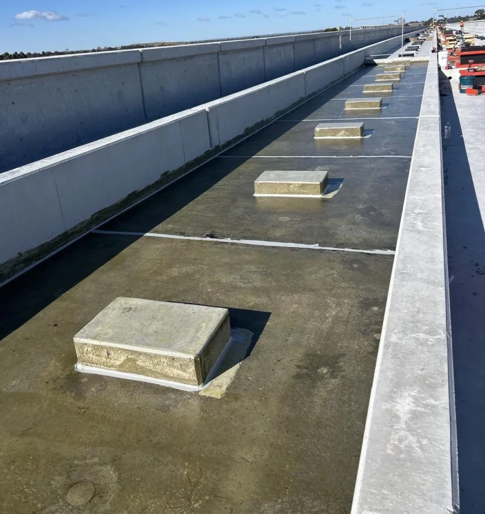A long concrete roadway with rectangular concrete blocks spaced evenly along its surface, bordered by concrete barriers on both sides, under a blue sky. Construction materials are visible to the right.