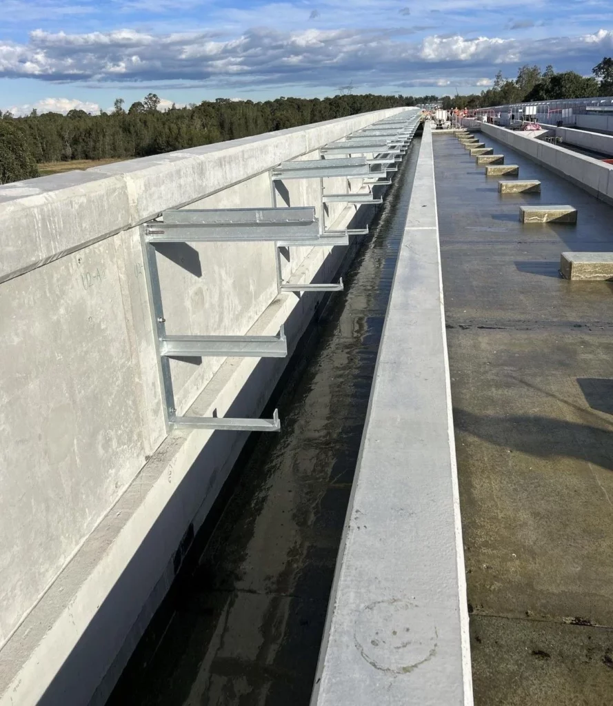 A long concrete bridge under construction features metal brackets extending from the side. The bridge surface is wet, and construction materials are visible. Trees and clouds appear in the background.