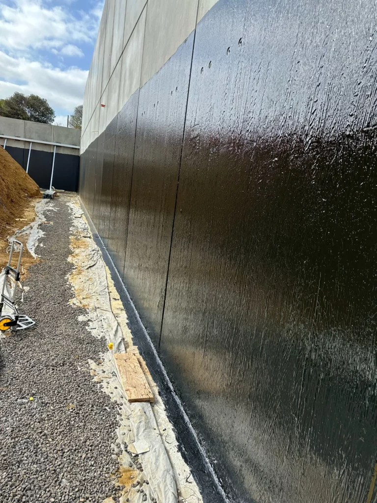 A concrete wall coated with black waterproofing material, gravel and plastic sheeting along its base, construction tools nearby, under a partly cloudy sky.