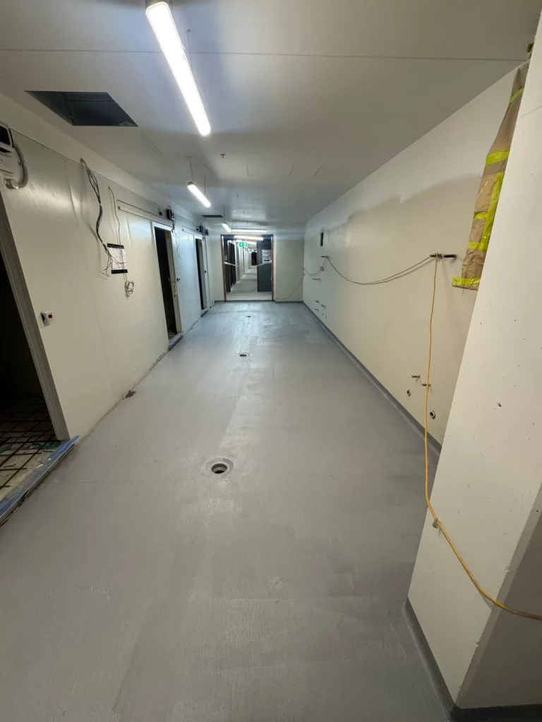 A hallway under construction with exposed wires, unfinished doorways, and a gray floor. Ceiling lights are on and some construction materials are visible to the side.