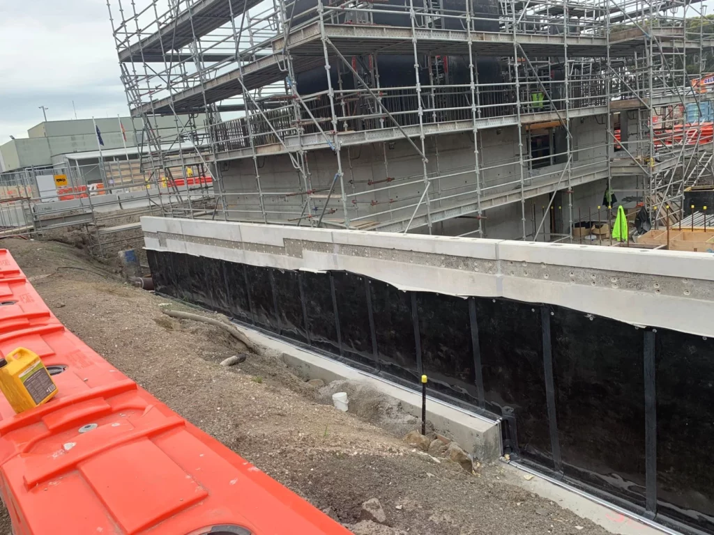 A construction site with scaffolding around a multi-story building. In the foreground, there are orange barriers, exposed soil, and a partially finished concrete wall lined with black waterproofing material.