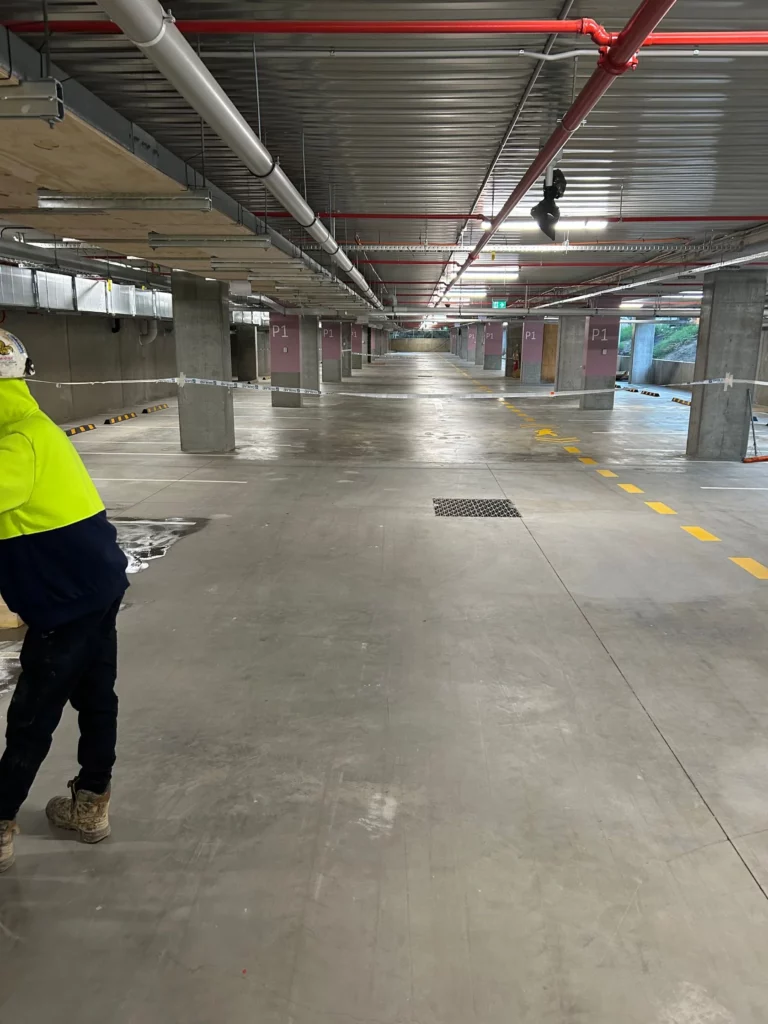 A worker in a hi-vis jacket stands in a mostly empty underground parking garage with concrete floors, pipes on the ceiling, and yellow striped markings on the right side.