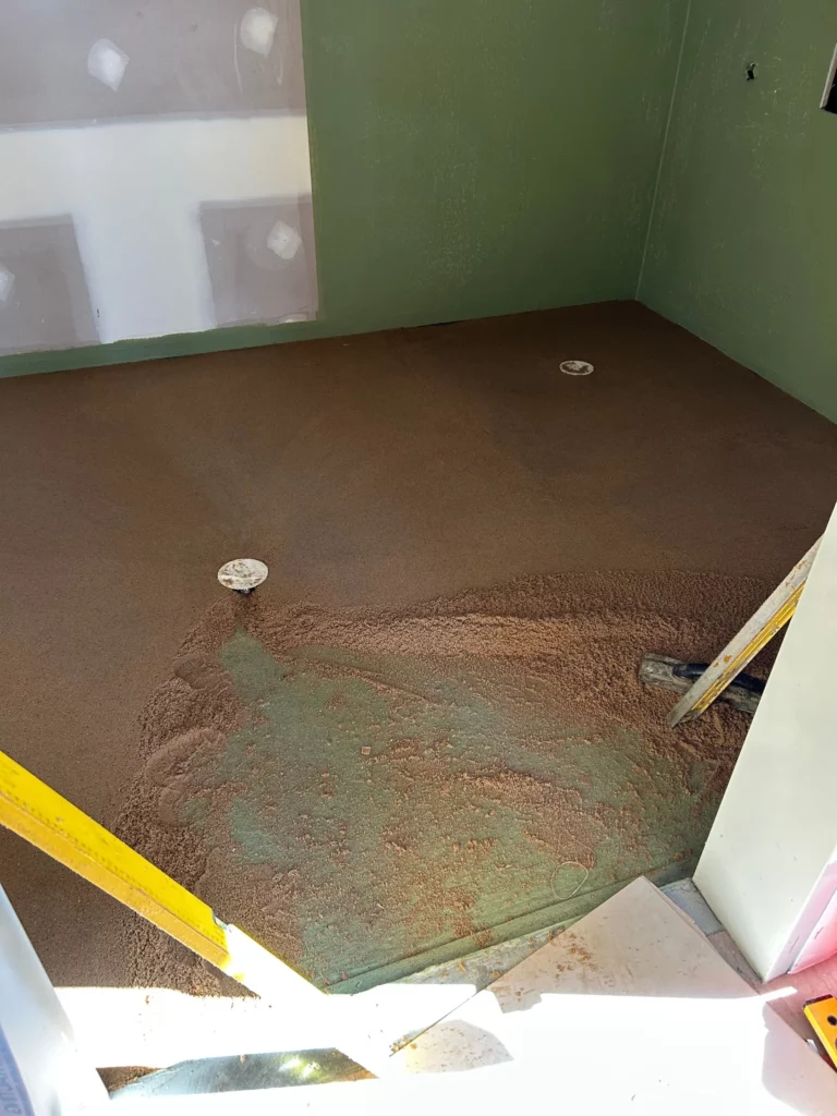 A room under construction with green walls, drywall, and a brown, freshly leveled floor. Two white plumbing pipes are on the floor, and a yellow leveling tool rests against the doorway.