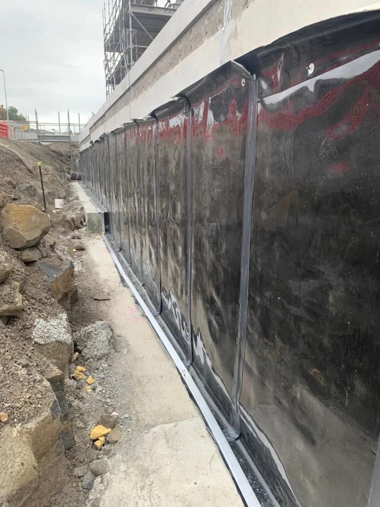 A concrete wall with black waterproofing membrane installed, surrounded by rocks and construction debris at an outdoor building site. Scaffolding and barriers are visible in the background.