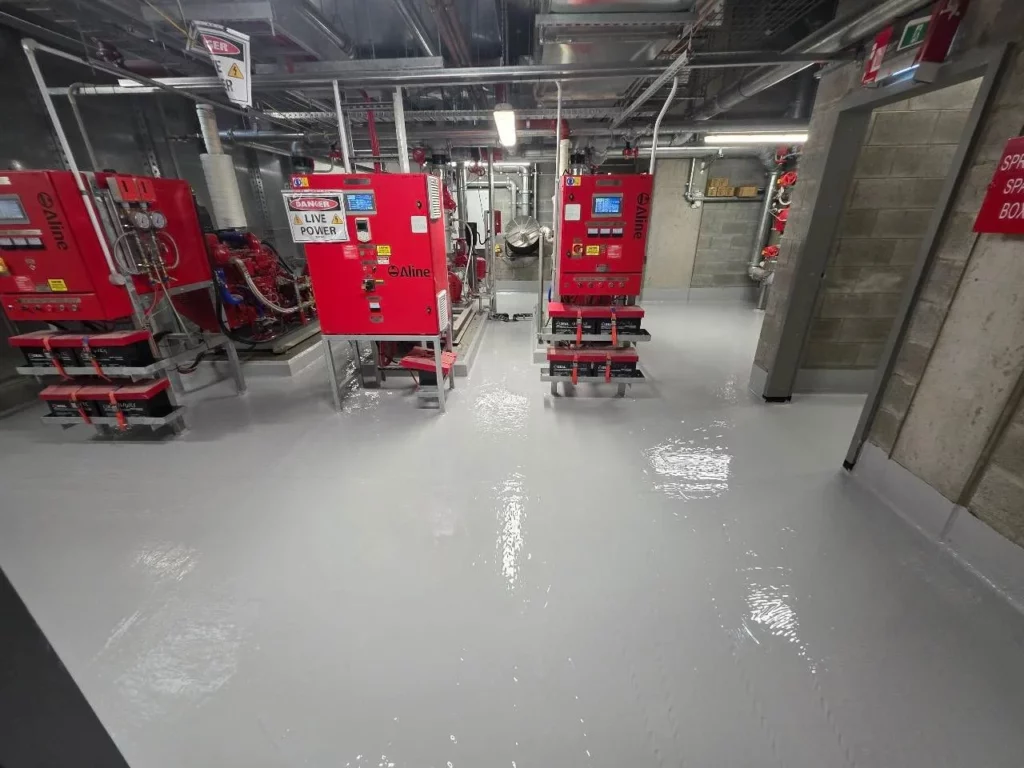 A room with a shiny, freshly coated floor, red industrial equipment, pipes, and exposed ceiling ducts. Signs and labels are visible on the machinery and walls.