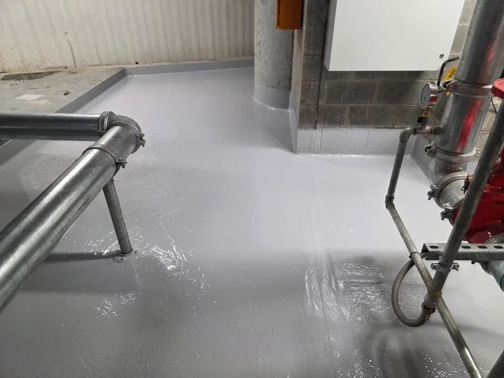 Industrial room with freshly applied, glossy gray floor coating, metal pipes and equipment visible, and a concrete pillar and block wall in the background.