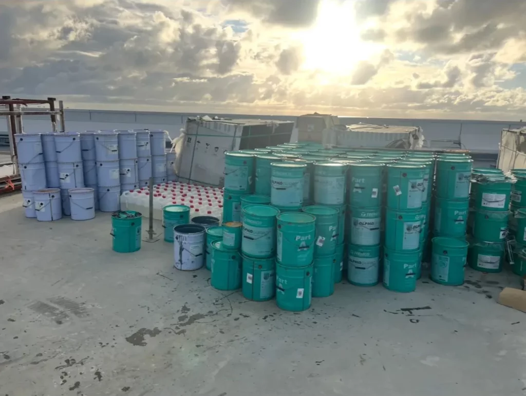 Several stacks of teal and white industrial barrels and containers are arranged on a concrete rooftop under a cloudy sky with sunlight peeking through.
